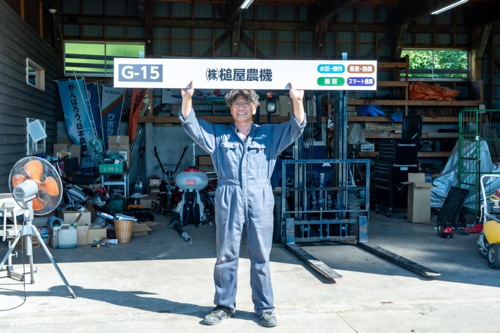 槌屋農機代表・中島慎也さんが屋外作業場で看板を掲げて笑顔を見せる様子。背後には農機具や工具が並び、活気ある現場の雰囲気を伝える。