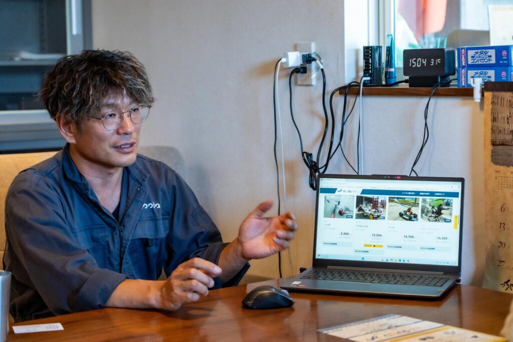 槌屋農機・中島代表がNRNSの仕組みについて説明する様子。パソコン画面には農機レンタル一覧が表示され、システムの具体的な利用画面が確認できる。