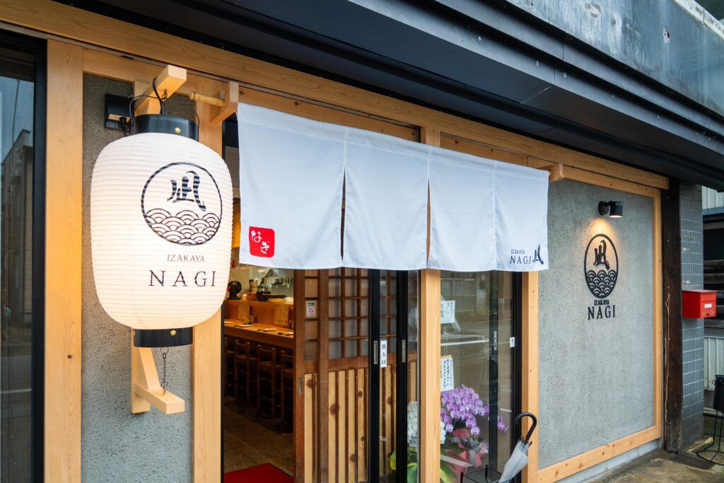 白い暖簾が印象的な「居酒屋 凪」の外観。木製の扉と温かみある照明が迎えてくれる。
