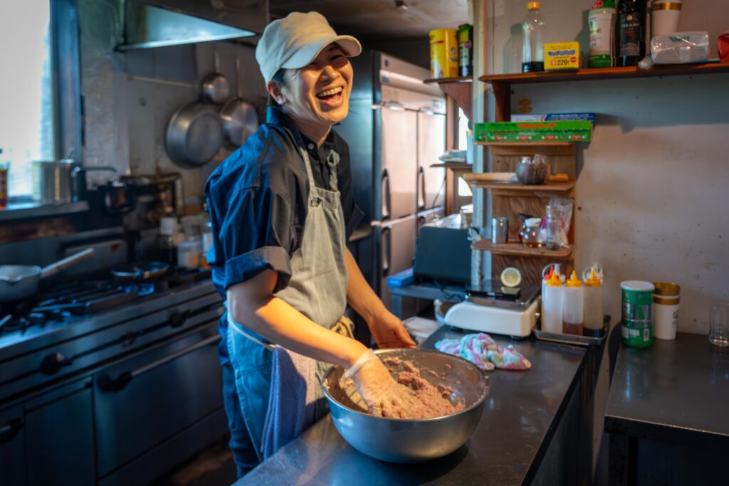 調理中の悠さんが笑顔で写るキッチン風景。たかきびハンバーグの仕込みをしている様子。