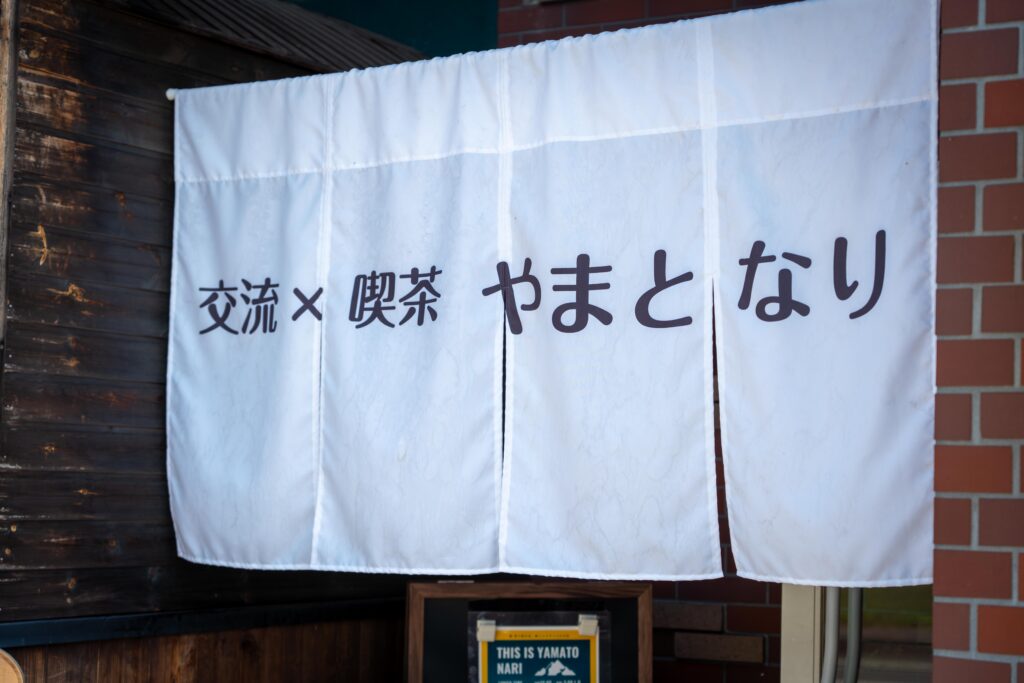 「交流×喫茶 やまとなり」と書かれた白いのれん。地域に開かれた場所であることを象徴する。
