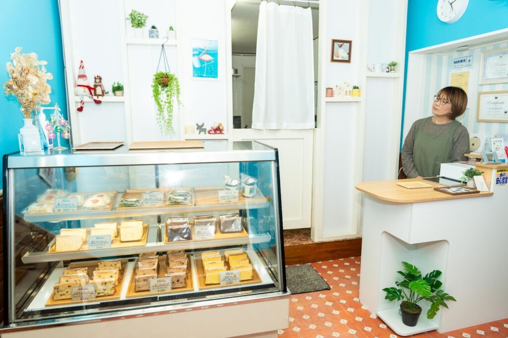 店内のショーケースに並ぶ米粉シフォンケーキと生米ロールケーキ。オーナーがカウンターに立ち、壁には食品衛生責任者などの資格証書が飾られている。