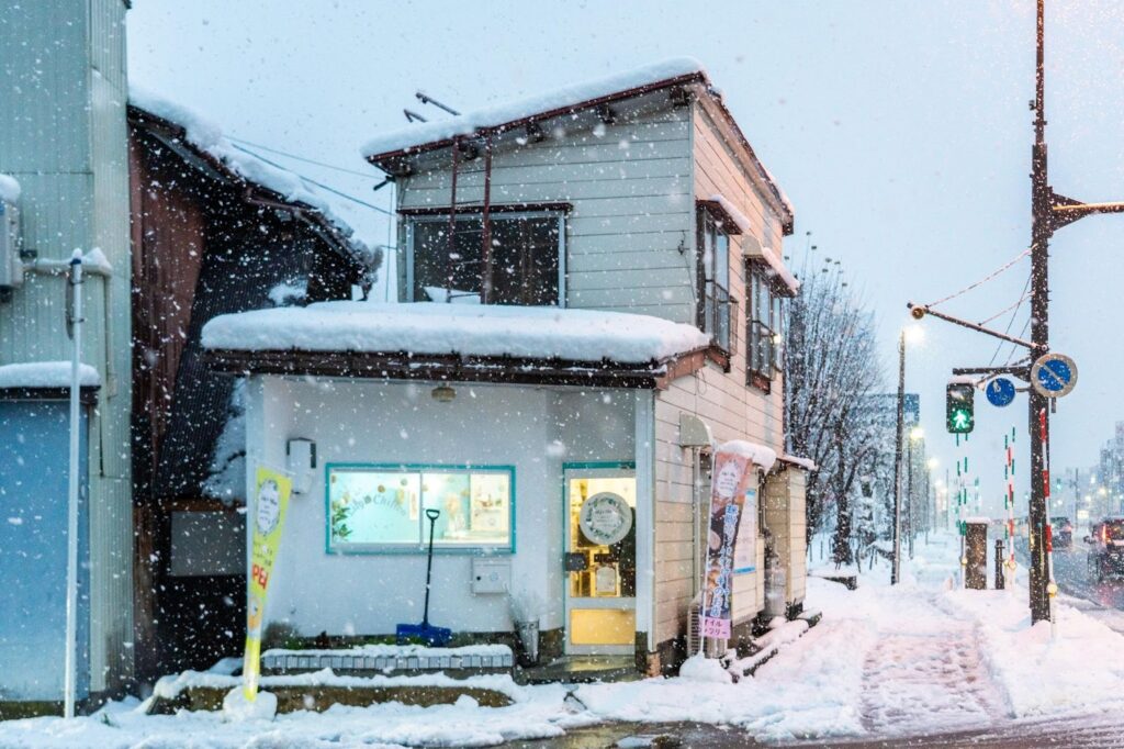 雪が降る南魚沼市六日町にあるLily's Chiffonの店舗外観。雪景色の中に温かい明かりが灯る、白い壁と青い窓枠の2階建ての小さなお店。